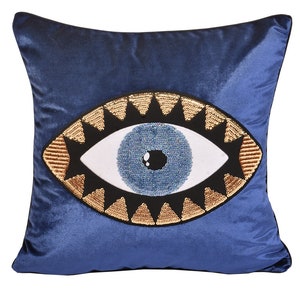 May include: Blue velvet pillow with an embroidered evil eye design. The eye is surrounded by gold and black sequins.