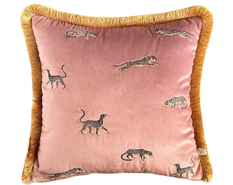 Puede incluir: Una almohada de terciopelo rosa con un borde de flecos dorados presenta un patrón bordado de leopardos negros y blancos.