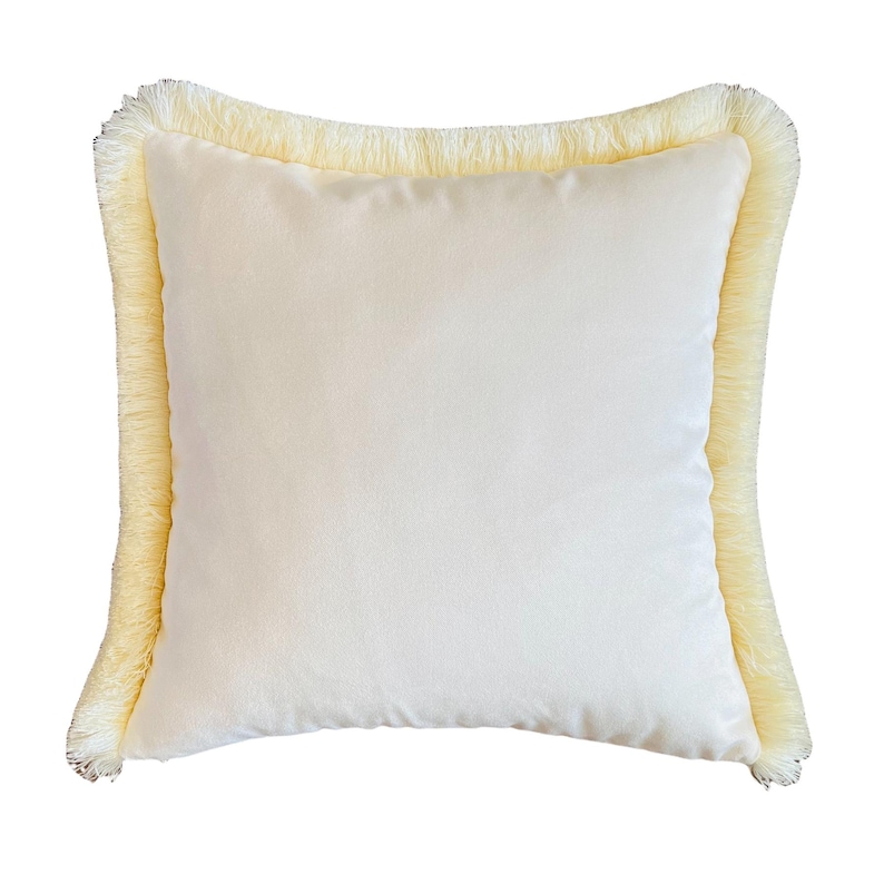 May include: A cream-colored pillow with a fringe border. The pillow is made of a soft, velvety fabric.