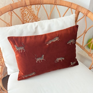 Puede incluir: Un cojín rectangular de terciopelo color óxido con guepardos bordados en varias poses. El cojín está colocado sobre un cojín blanco, con una silla de mimbre en el fondo. Los guepardos son en tonos marrones y negros.