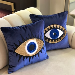 May include: Two blue velvet throw pillows with gold and blue evil eye designs. The pillows are on a light beige couch.