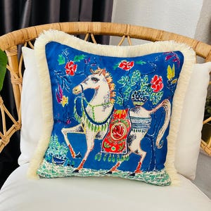 May include: A blue throw pillow with a white horse design and colorful floral accents. The pillow has a white fringe border.