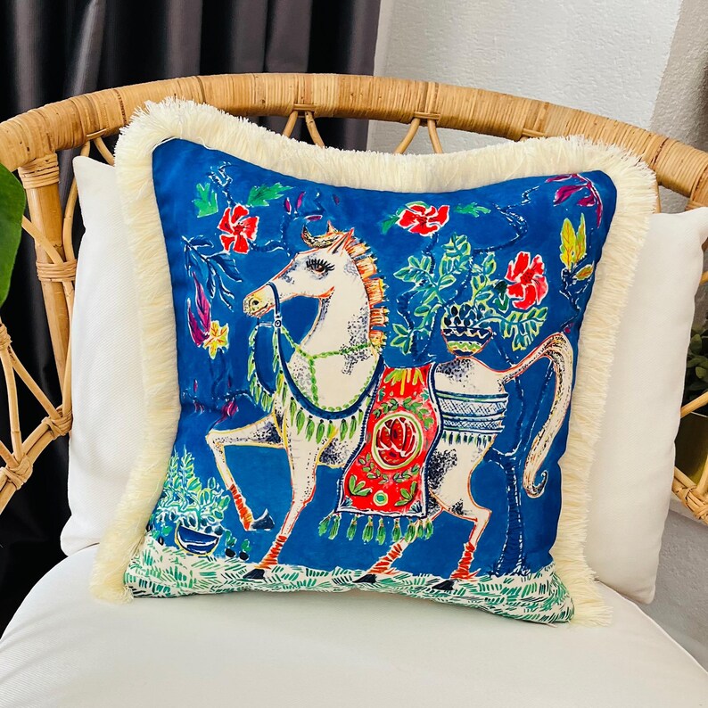 May include: A blue throw pillow with a white horse design and colorful floral accents. The pillow has a white fringe border.