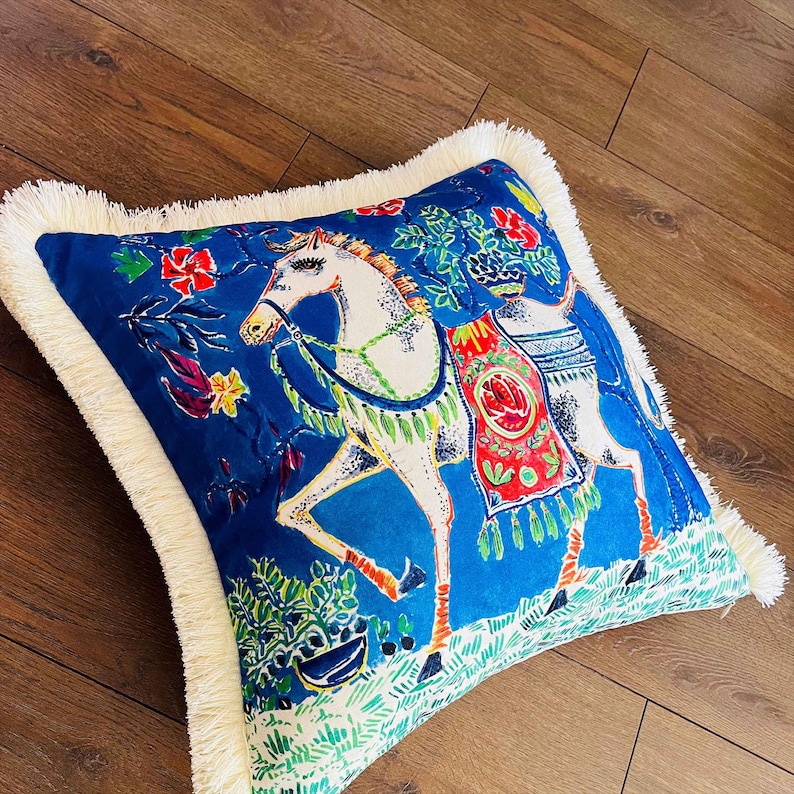 May include: A blue throw pillow with a white fringe border features a colorful illustration of a white horse with a red and green saddle. The horse is surrounded by green and blue floral designs.