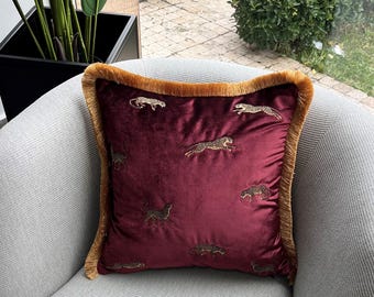 Puede incluir: Una almohada de terciopelo burdeos con un borde de flecos dorados presenta un patrón repetido de leopardos bordados.