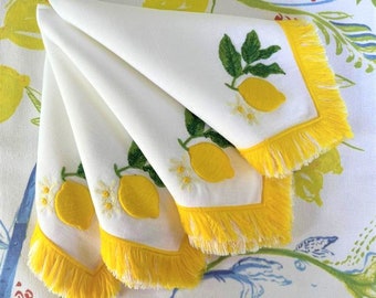 May include: Set of four white linen napkins with yellow fringe and embroidered lemons. The napkins are folded and arranged on a white tablecloth with a lemon print.