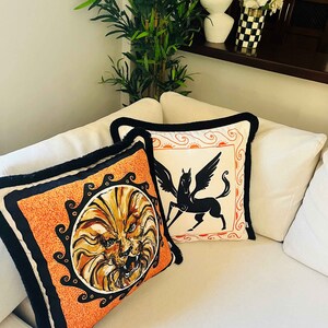 May include: Two decorative throw pillows with black trim. One pillow features a lion's head in a circle on an orange background. The other pillow features a black silhouette of a winged horse on a white background.