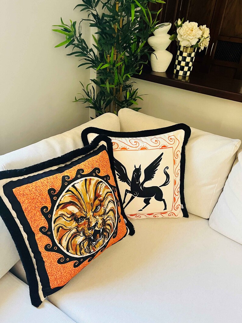 May include: Two decorative throw pillows with black trim. One pillow features a lion's head in a circle on an orange background. The other pillow features a black silhouette of a winged horse on a white background.