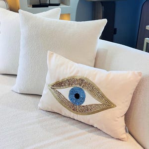 May include: A pink velvet throw pillow with a large blue and gold evil eye design. The eye is made of sequins and beads.