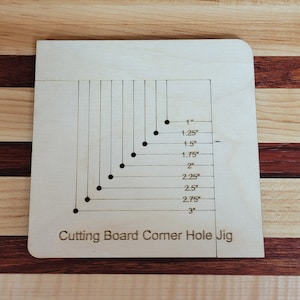 May include: A wooden cutting board corner hole jig with markings for 1 inch to 3 inches in increments of 0.25 inches. The jig is made of light wood and has a natural finish.