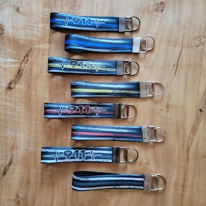 May include: Set of ten keychains with different designs featuring the number 911 and a heartbeat graphic. The keychains are made of black and white striped fabric with different colored accents.