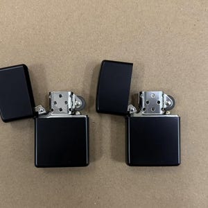 May include: Two black lighters with open lids, showcasing their metallic interiors. The lighters are rectangular with rounded corners, set against a neutral background. The lids are hinged and open to reveal the lighter's mechanism.