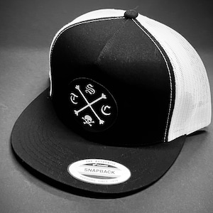 May include: Black and white trucker hat with a skull and crossbones logo and the text "A.S.C."  The hat has a white mesh back and a black front with a black "The Authentic Yupoong Snapback" label.