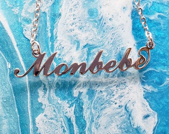 May include: A silver necklace with the name "Monbebe" spelled out in cursive script. The necklace is on a blue and white background.
