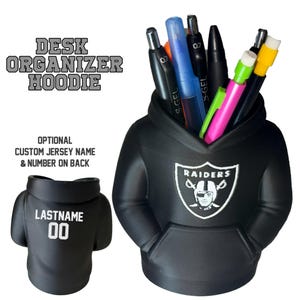 May include: A black desk organiser in the shape of a hoodie, with the Raiders logo. The hoodie holds pens and pencils of various colours. The text "DESK ORGANIZER HOODIE" is above the organiser. The back of the hoodie has the text "LASTNAME" and the number "00".