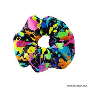 Pode incluir: Um scrunchie preto com um design de respingos de tinta colorida. O scrunchie apresenta cores brilhantes como rosa, amarelo, verde, azul e laranja.