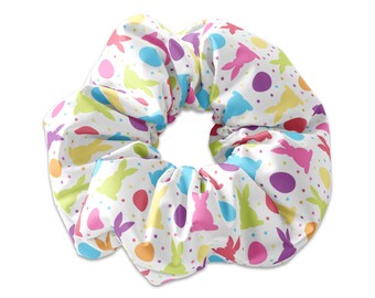 May include: A white scrunchie with a colourful Easter bunny and egg pattern. The scrunchie is made of soft, stretchy fabric and is perfect for holding back hair.