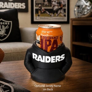 May include: A black hoodie-shaped can cooler with the word "RAIDERS" in white. An orange can of "BLOOD ORANGE IPA" beer sits inside. A football and a pillow with the Raiders logo are in the background.
