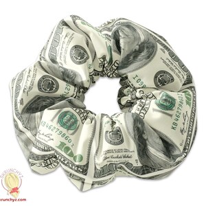 May include: A white scrunchie with a pattern of US dollar bills. The scrunchie is made of fabric and has a gathered design. The bills feature the portrait of George Washington and the text "ONE HUNDRED DOLLARS".