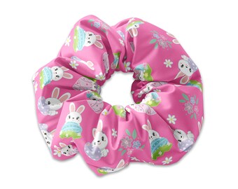 May include: A pink scrunchie featuring a repeating pattern of Easter bunnies, eggs, and flowers. The bunnies have blue and green accents. This hair accessory is made of soft fabric.