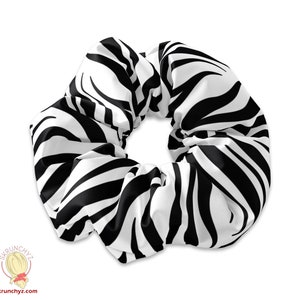 Puede incluir: Un gran coletero con un estampado de cebra en blanco y negro. El accesorio para el cabello está hecho de una tela suave, posiblemente satén. El coletero está diseñado para sujetar el cabello en una cola de caballo o moño.