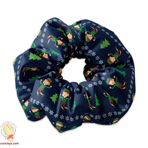 Könnte beinhalten: Ein marineblauer Scrunchie mit einem sich wiederholenden Muster aus Elfen, die um Weihnachtsbäume tanzen. Die Elfen tragen grüne und rote Outfits und weiße Hüte. Der Hintergrund ist dunkelblau mit weißen Schneeflocken.