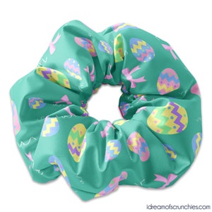 May include: A teal blue scrunchie with a repeating pattern of pastel pink, yellow, and purple Easter eggs. The eggs have a variety of designs, including stripes, polka dots, and zigzags.