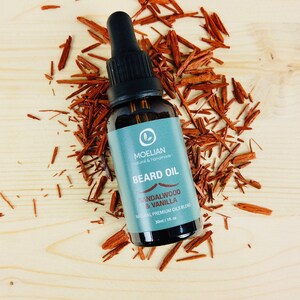 May include: A dark brown glass bottle with a dropper top filled with beard oil. The bottle has a blue label with white text that reads "Moellan Natural & Handmade Beard Oil Sandalwood & Vanilla Natural Premium Oils Blend 30ml / 1 fl oz". The bottle is surrounded by red wood shavings on a light wood surface.