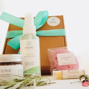 May include: A collection of natural skincare products, including body oil, face scrub, and a face and body toner. The products are packaged with a gift box tied with a teal ribbon, a lavender soap, and lip balm. The products are arranged on a white surface.