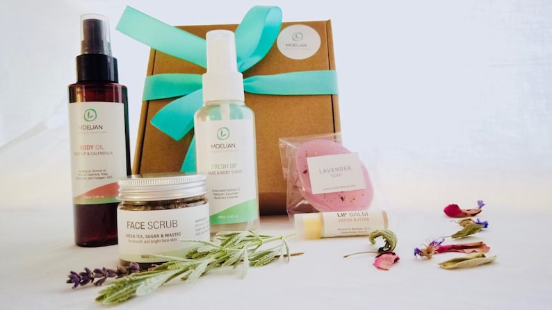 May include: A gift set of skincare products, including body oil, face scrub, face and body toner, lavender soap, and lip balm. The products are in clear and brown bottles and jars, with a brown box tied with a teal ribbon. Dried flowers are scattered around.