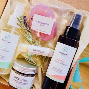 May include: A gift box containing skincare products. Includes a bottle of face and body toner, a jar of face scrub, a bottle of body oil, a lip balm, and a lavender soap. The products are arranged with sprigs of herbs.