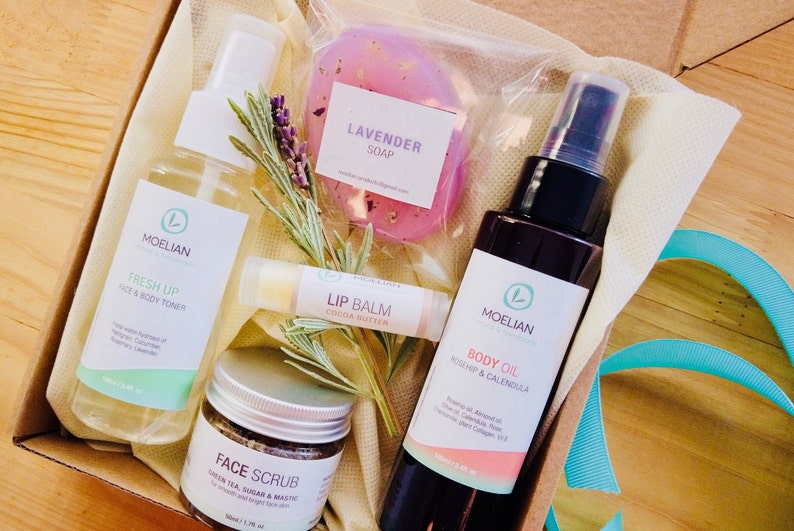 May include: A gift box containing skincare products. Includes a bottle of face and body toner, a jar of face scrub, a bottle of body oil, a lip balm, and a lavender soap. The products are arranged with sprigs of herbs.