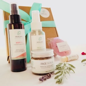 May include: A gift set of natural skincare products, including body oil, face scrub, and a lavender-scented bath bomb. The set is packaged in a brown box tied with a teal ribbon. Additional items include a lip balm and a fresh up spray.