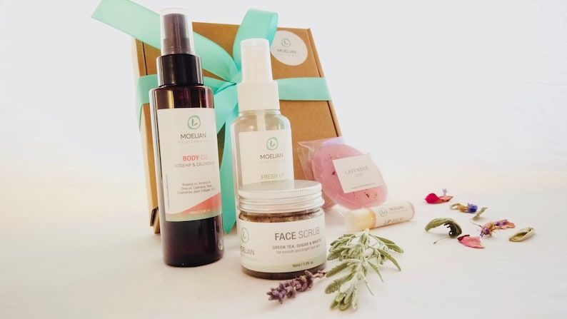 May include: A gift set of natural skincare products, including body oil, face scrub, and a lavender-scented bath bomb. The set is packaged in a brown box tied with a teal ribbon. Additional items include a lip balm and a fresh up spray.