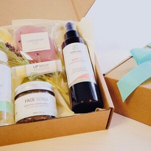 May include: A gift box filled with beauty products, including a lavender soap, lip balm, face scrub, and body oil. The box is brown with a light blue ribbon on top. The products are labeled with the brand name "Moelian".