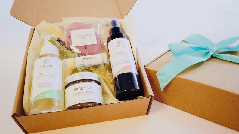 May include: A gift box filled with beauty products, including a lavender soap, lip balm, face scrub, and body oil. The box is brown with a light blue ribbon on top. The products are labeled with the brand name "Moelian".