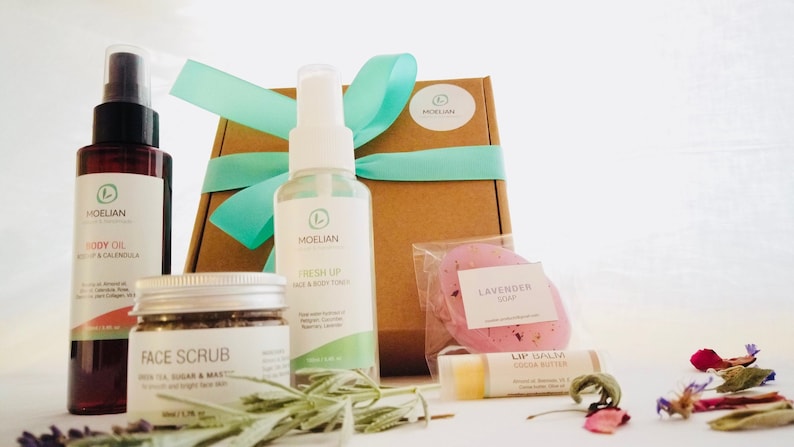May include: A collection of natural skincare products, including body oil, face scrub, and a face and body toner. The products are packaged with a gift box tied with a teal ribbon, a lavender soap, and lip balm. The products are arranged on a white surface.