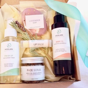 May include: A gift box filled with beauty products, including a lavender soap, lip balm, face scrub, body oil, and face and body toner. The products are arranged with sprigs of rosemary and a light blue ribbon.