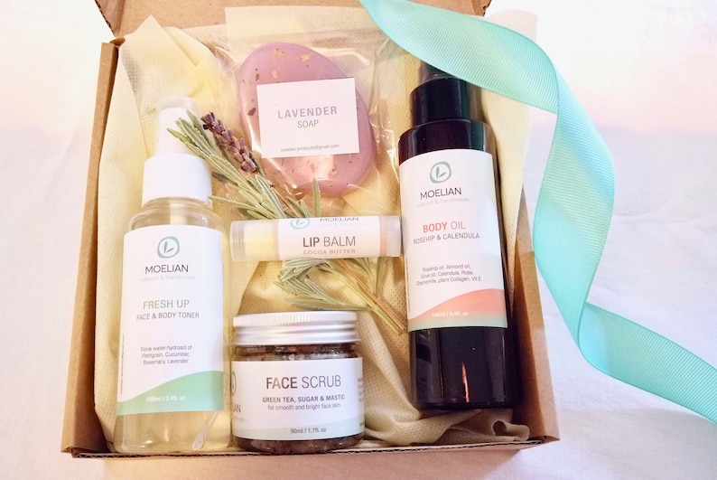 May include: A gift box filled with beauty products, including a lavender soap, lip balm, face scrub, body oil, and face and body toner. The products are arranged with sprigs of rosemary and a light blue ribbon.