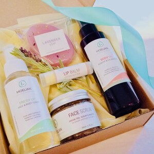 May include: A gift box filled with natural skincare products. The box contains a lavender soap, body oil, face scrub, lip balm, and face and body toner. The products are packaged with dried lavender and a light blue ribbon.