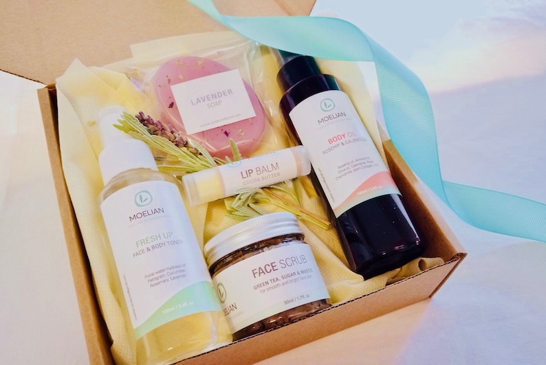 May include: A gift box filled with natural skincare products. The box contains a lavender soap, body oil, face scrub, lip balm, and face and body toner. The products are packaged with dried lavender and a light blue ribbon.
