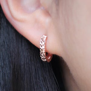 May include: Rose gold hoop earring with a textured, geometric design. The earring is a small, circular shape and is worn on the ear. The earring is a warm, metallic color.