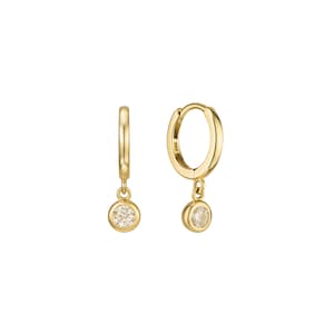 May include: A pair of gold hoop earrings, each featuring a single round, bezel-set clear gemstone charm dangling from the bottom. The metal has a polished finish, and the hoops are designed with a hinged closure mechanism for secure wear.