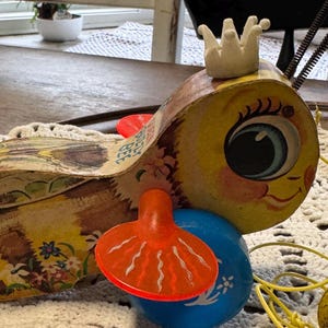 May include: Vintage wooden pull toy shaped like a bird with a crown. The bird has a yellow body, blue and orange accents, and a large eye. The toy is on a white crocheted doily.