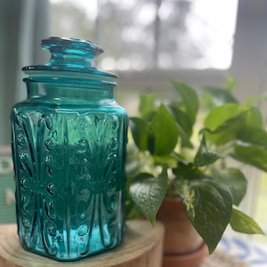 Könnte beinhalten: Eine tealblaue Glasdose mit Deckel und einem dekorativen, geprägten Muster. Die Dose steht auf einem Holzsockel.