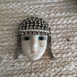Peut inclure: Broche vintage en forme de masque avec un visage en porcelaine blanche, des yeux noirs et une bouche rose. Le masque est orné de strass et de fausses perles, avec des mèches de strass de chaque côté. Un seul strass rouge est sur la joue.