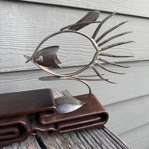 May include: A handcrafted metal fish sculpture with a spoon base, tail, and fins. The fish is silver-colored and sits on a dark brown wooden stand. The sculpture is displayed on a weathered wooden surface.