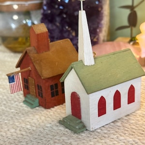 May include: Two miniature wooden buildings, a red schoolhouse with an American flag and a white church with a green roof and a white steeple. The buildings are on a white textured surface.