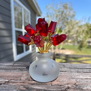 May include: A small frosted glass vase with a textured surface holds a bouquet of red glass flowers with gold stems.