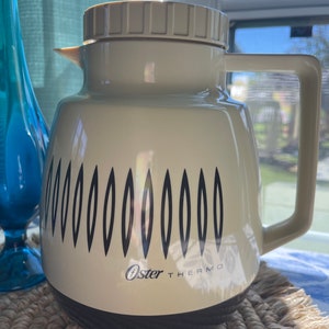 May include: A vintage white Oster Thermo carafe with a black and white geometric design. The carafe has a lid and a handle.